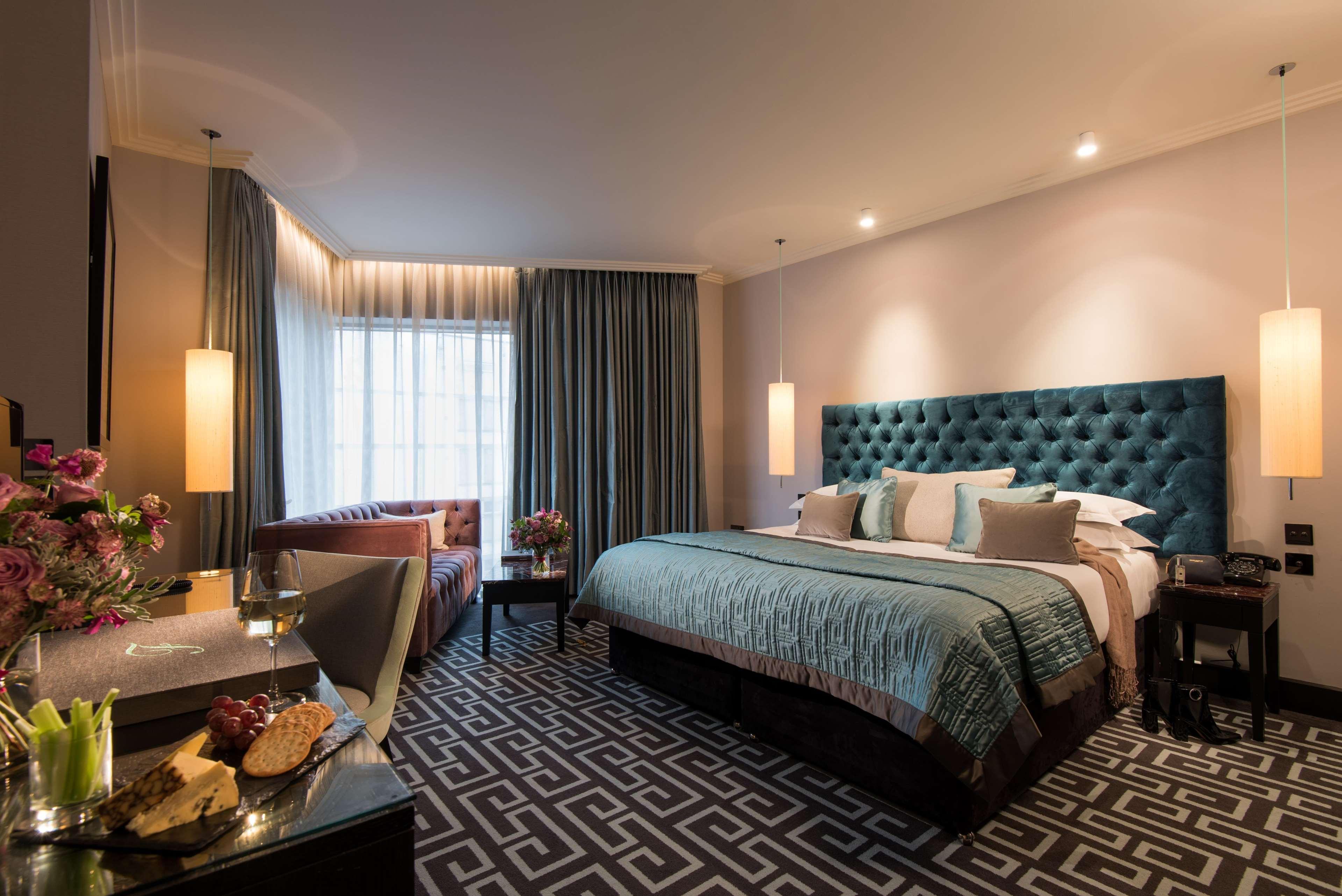 فندق ذا فيتزويليام بلفاست المظهر الخارجي الصورة The photo depicts a stylish hotel room. It features a large bed with a tufted teal headboard, flanked by two wall sconces providing soft lighting. The bedding is neatly arranged with accent pillows. To one side, there is a small seating area with a c