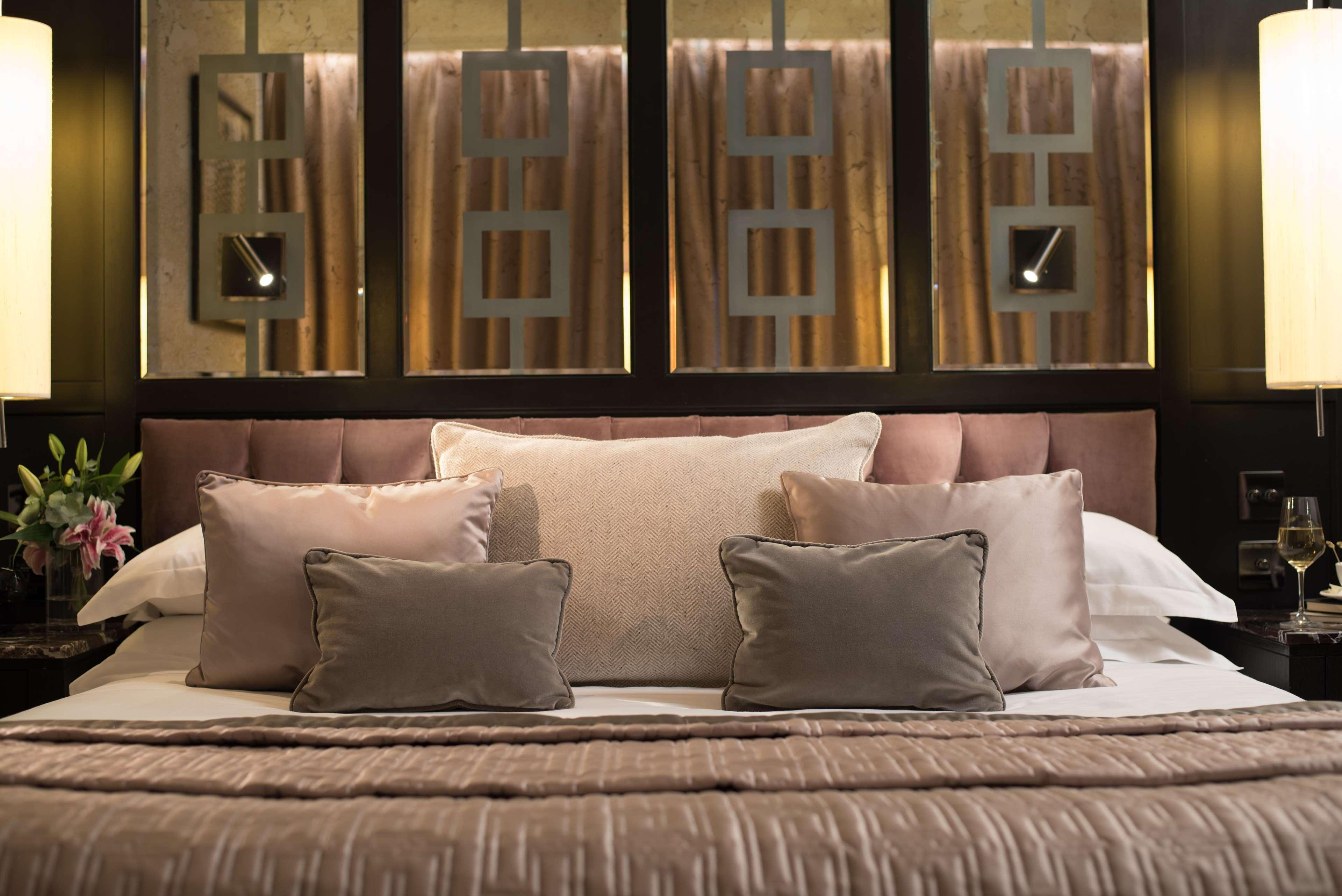 فندق ذا فيتزويليام بلفاست المظهر الخارجي الصورة The photo shows a beautifully designed bed in a stylish bedroom. The bed is adorned with a soft, plush headboard and layered with various decorative pillows in shades of beige and gray. The bedding appears luxurious, with a textured blanket. Behind t