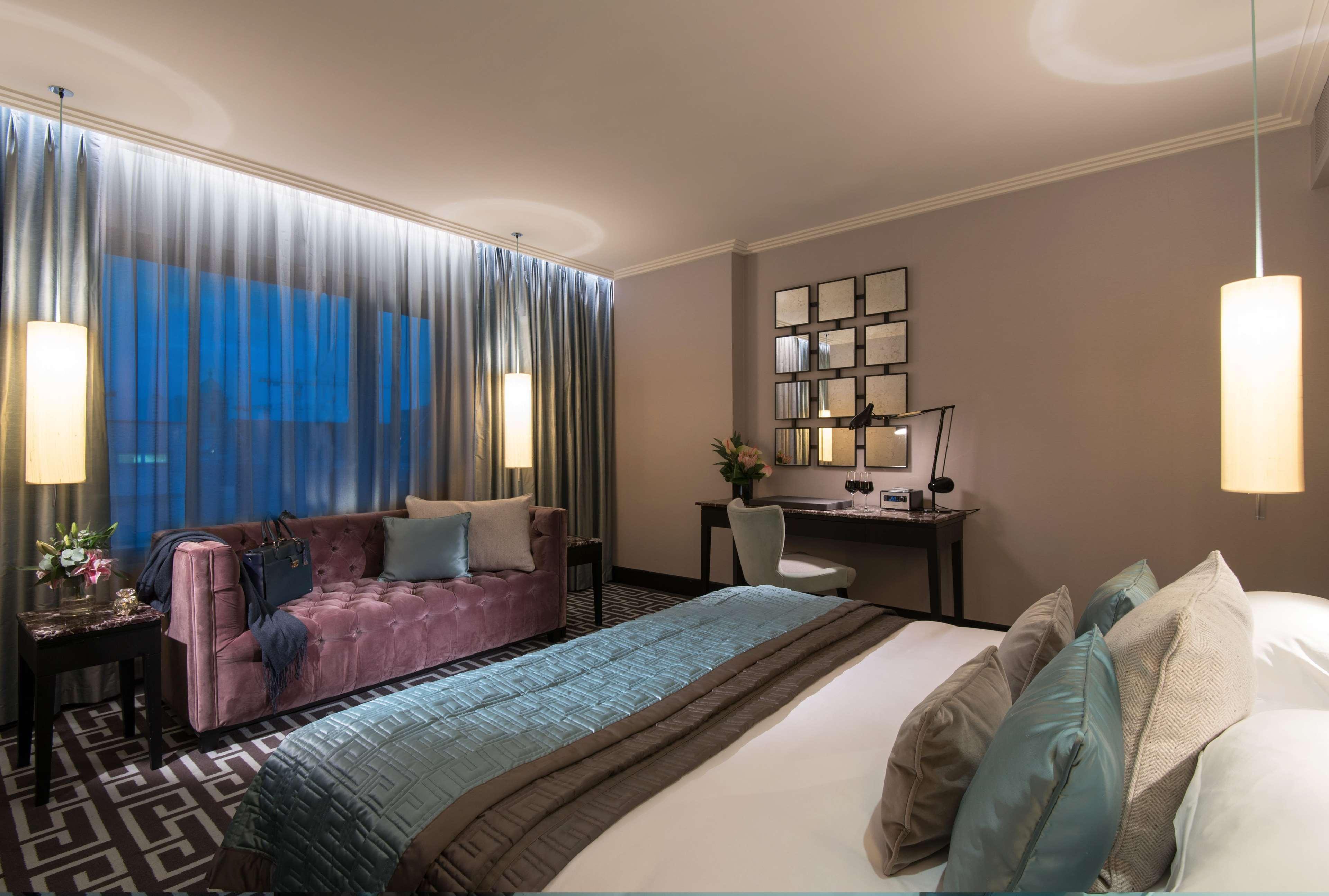 فندق ذا فيتزويليام بلفاست المظهر الخارجي الصورة The photo depicts a modern hotel room featuring a spacious layout. There is a large bed with decorative pillows in the foreground, covered with a stylish bedspread. To one side, a plush purple sofa adds a pop of color and comfort. A small desk and ch
