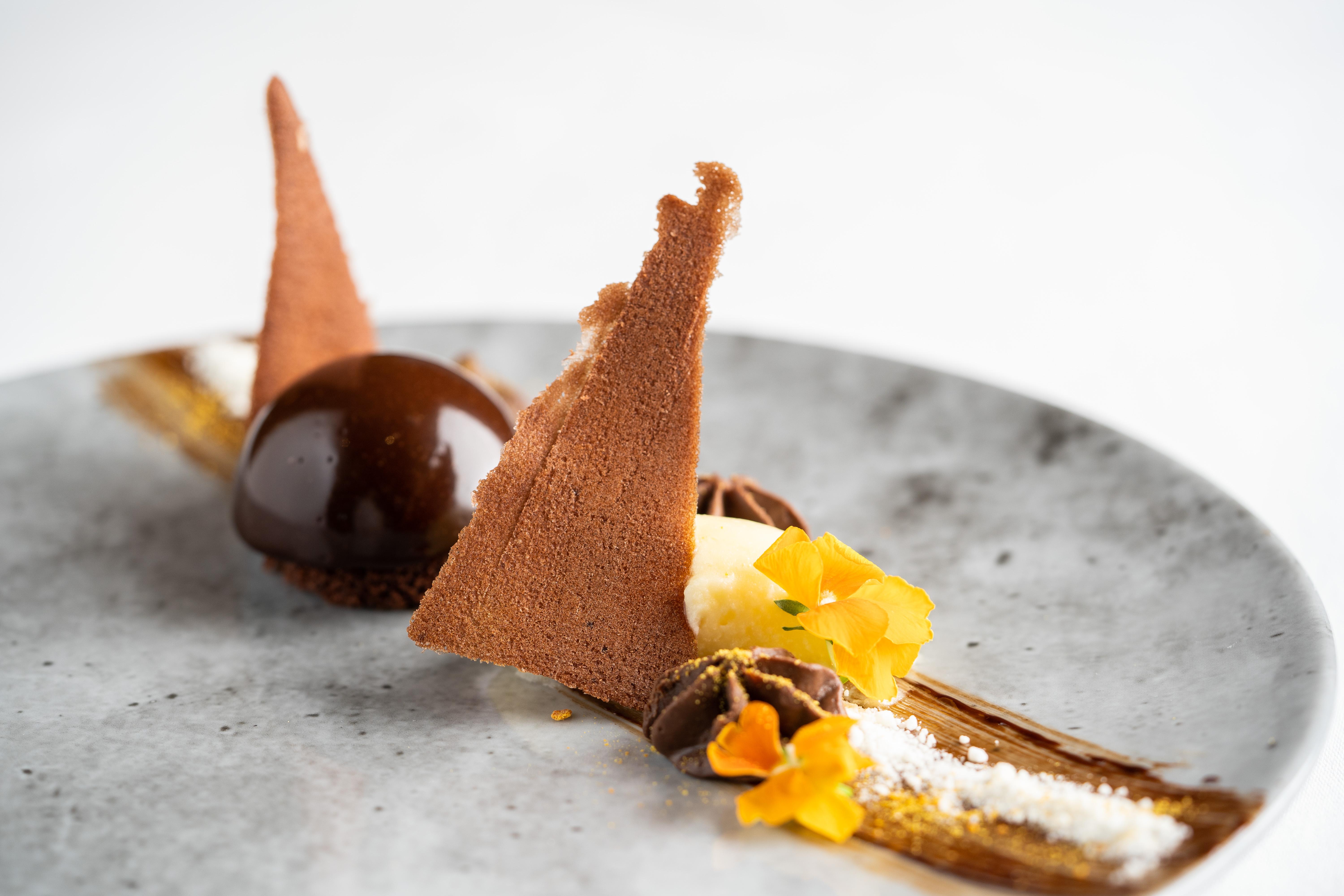 فندق ذا فيتزويليام بلفاست المظهر الخارجي الصورة The photo displays an artfully plated dessert. It features a glossy chocolate sphere, accompanied by various chocolate components, including triangular chocolate tuile pieces that resemble sails. The plate is adorned with delicate garnishes such as y