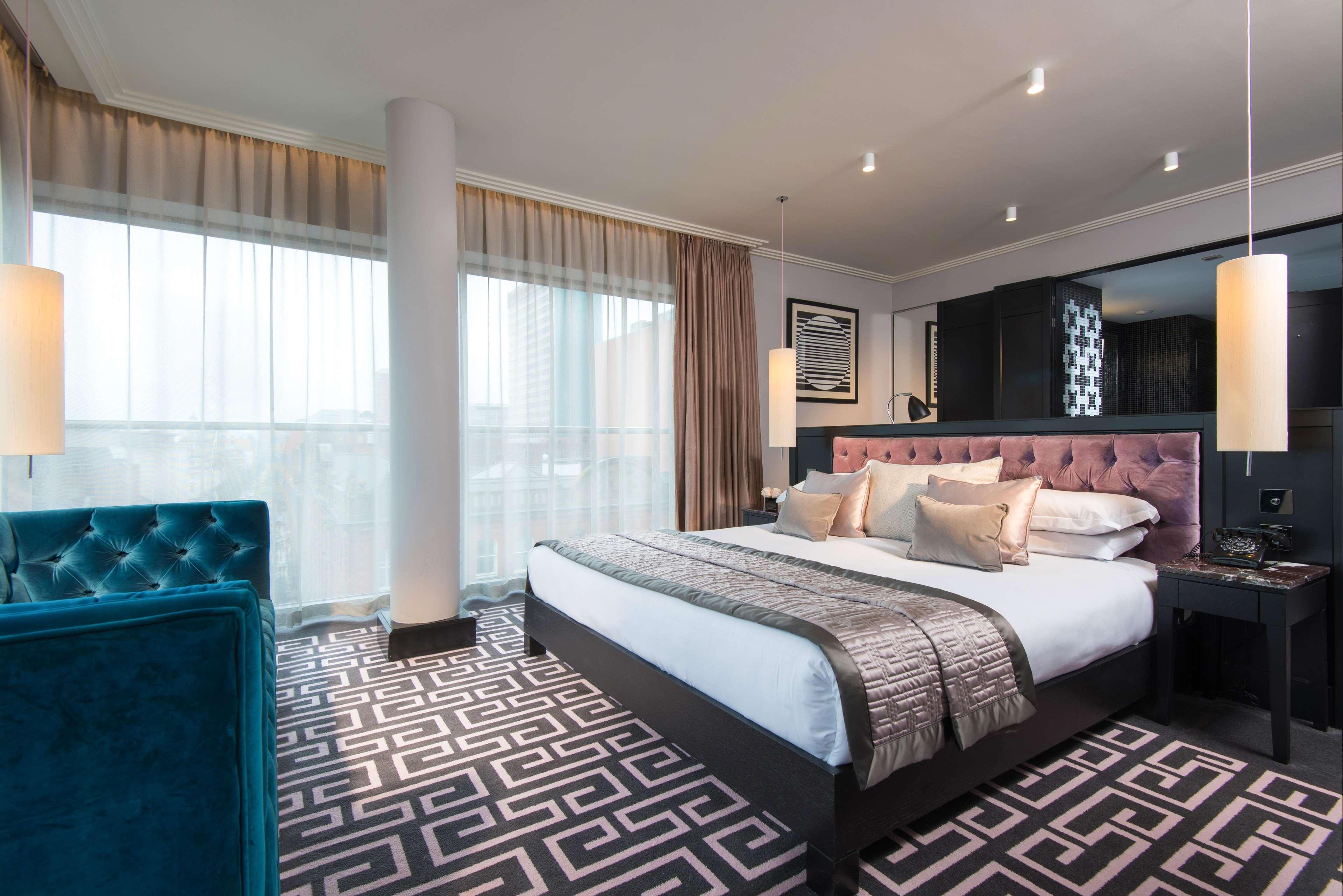 فندق ذا فيتزويليام بلفاست المظهر الخارجي الصورة The photo showcases a modern hotel room. It features a large bed with plush pillows and a stylish headboard. The room has large windows with sheer curtains, allowing natural light to filter in. The floor is covered with a patterned carpet that contra