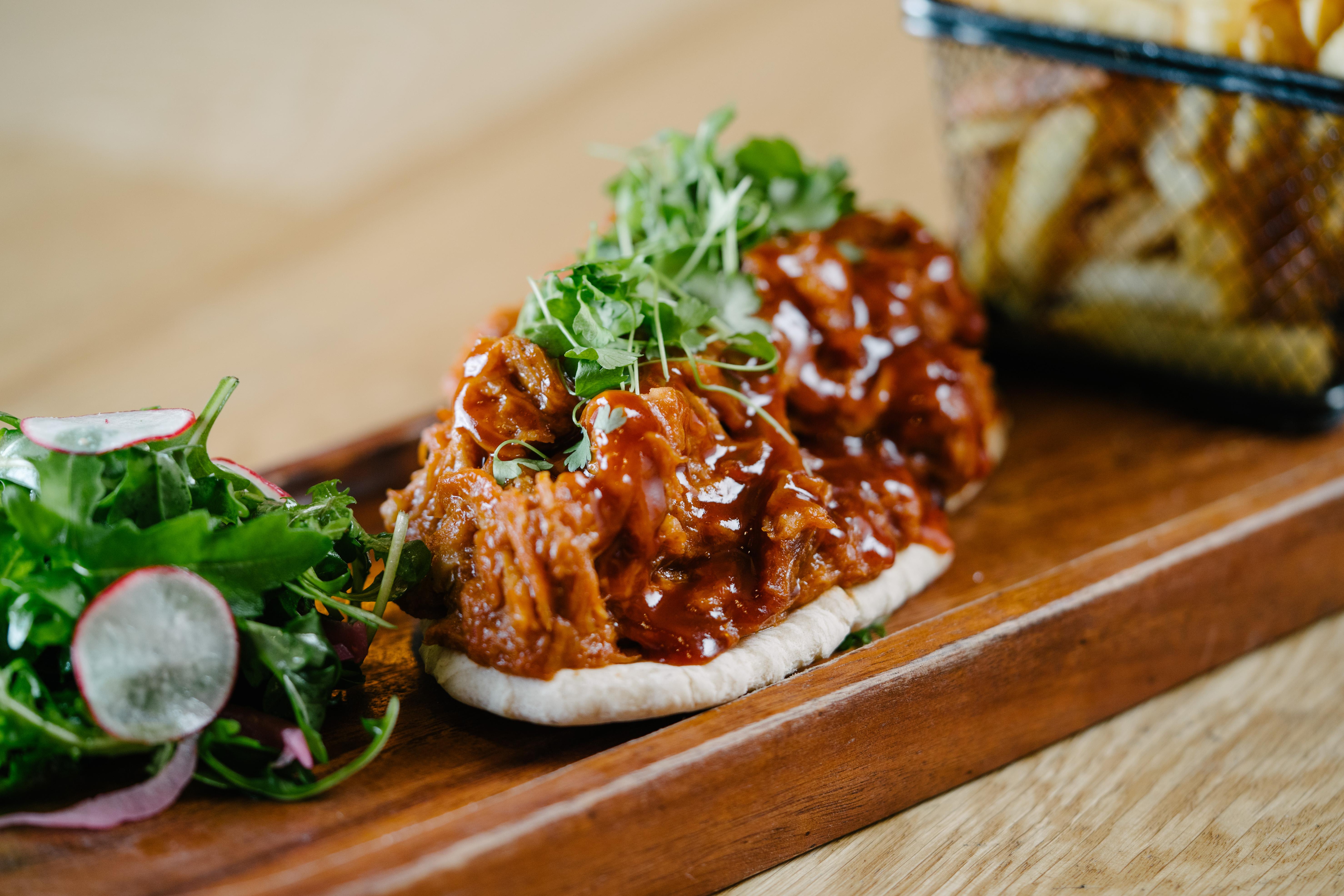 فندق ذا فيتزويليام بلفاست المظهر الخارجي الصورة The photo shows a wooden platter featuring two pieces of bread topped with pulled meat covered in a rich barbecue sauce. The dish is garnished with fresh greens, including sprouts. On the side, there is a small serving of mixed salad with radishes an